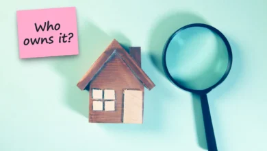 A small wooden house model next to a magnifying glass on a pale blue background, with a sticky note reading “Who owns it?”.