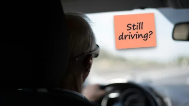 An older man with white hair seen from behind at the wheel of a car, with a sticky note reading “Still driving?”.