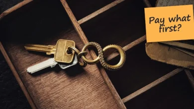 House keys in a wooden tray beside a stack of old letters, with a sticky note reading “Pay what first?”.