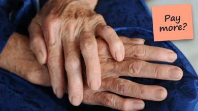 An older person's folded hands resting on blue fabric, with a sticky note reading “Pay more?”.
