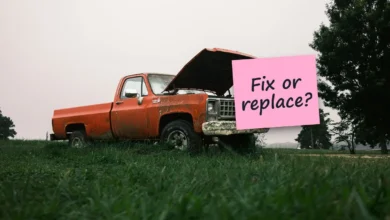An old orange pickup truck with its hood up, parked on grass, with a sticky note reading “Fix or replace?”.